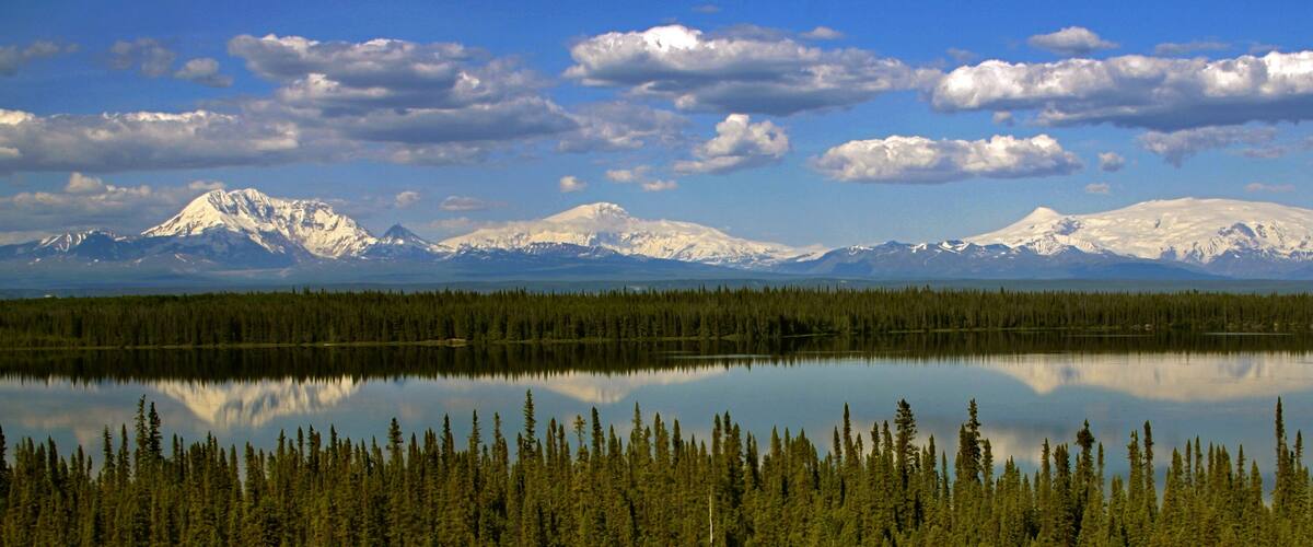 A calm lake reflecting snow-capped mountains and a dense forest.