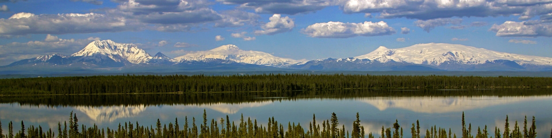 A calm lake reflecting snow-capped mountains and a dense forest.