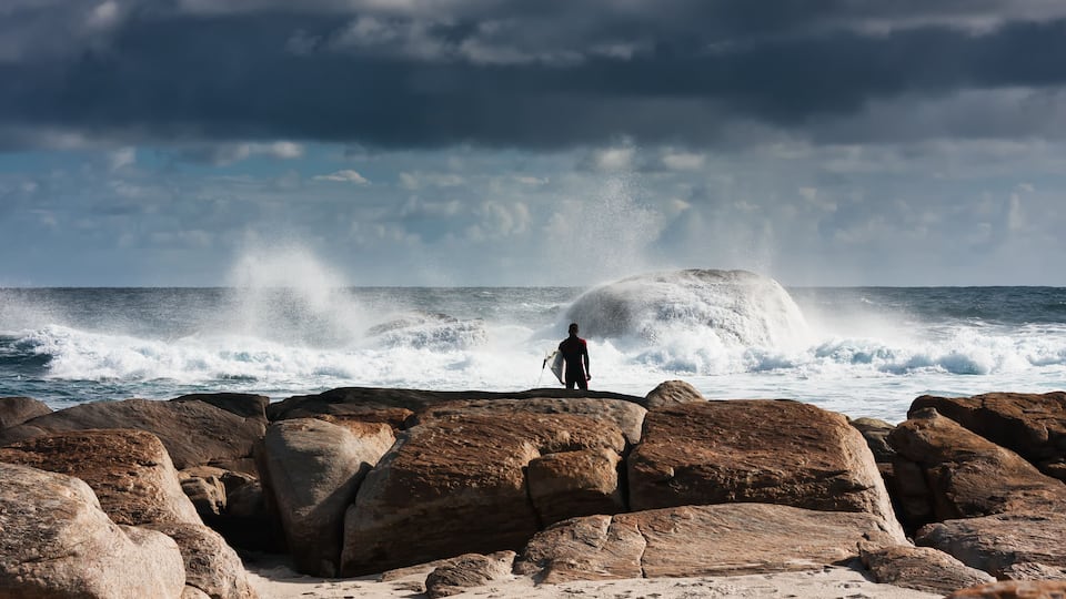 A surfer standing on rocks by the ocean with waves crashing.