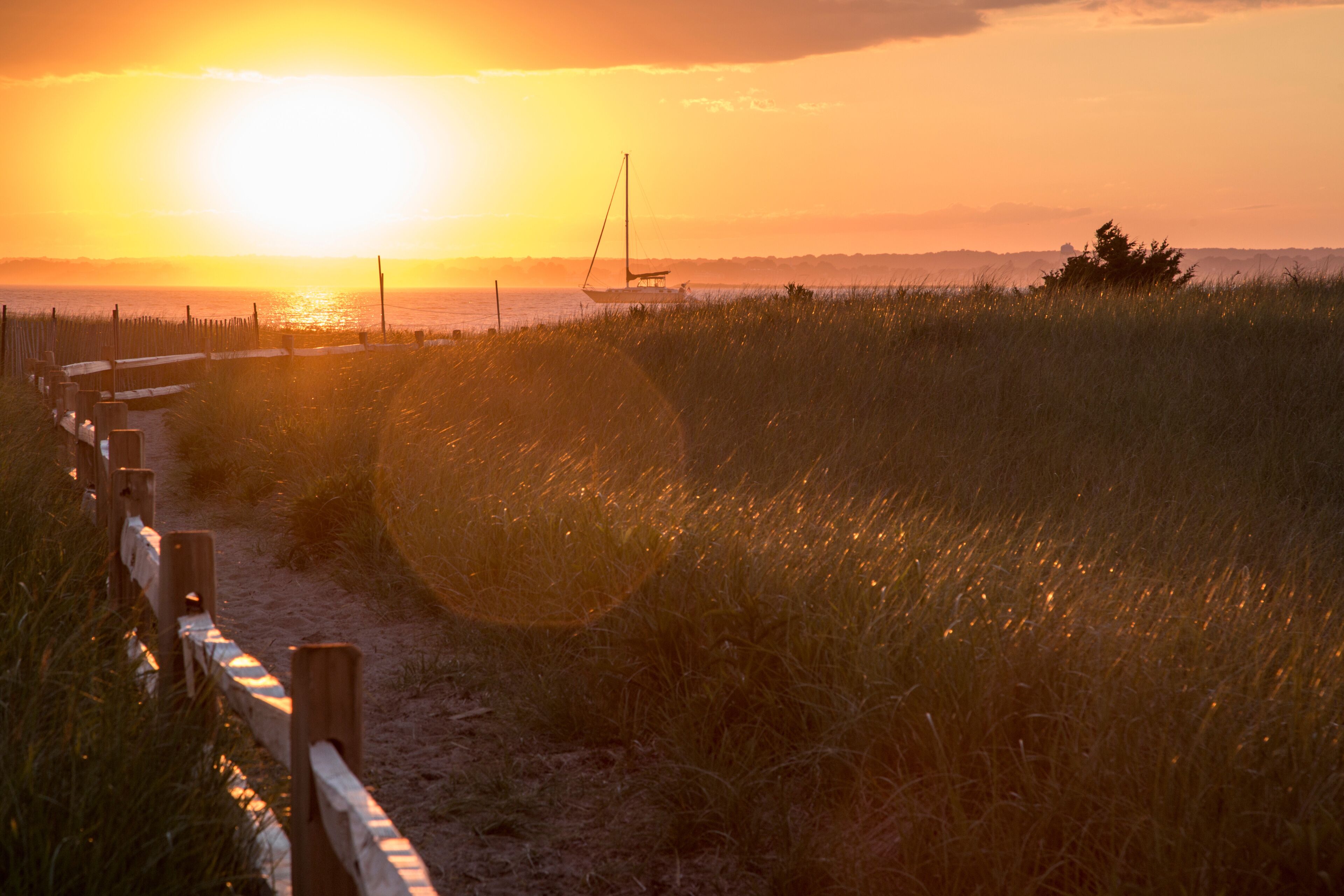 Beach path, wooden fence and sailboat at sunset at Napatree Point, Watch Hill, Westerly, Rhode Island, USA