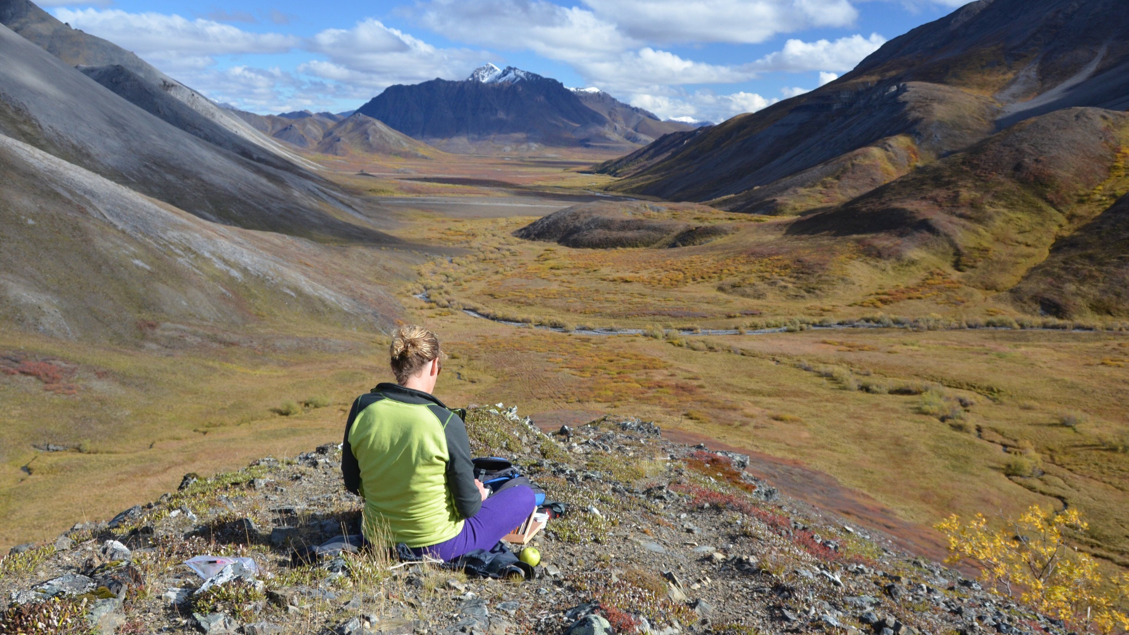 A person sitting on rocky terrain overlooking a valley with mountains in the background.