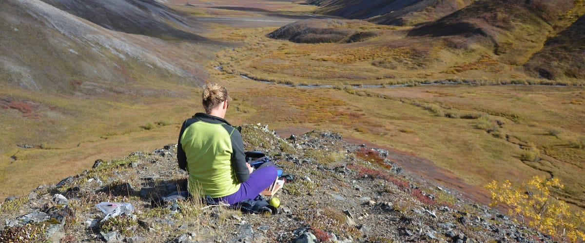 A person sitting on rocky terrain overlooking a valley with mountains in the background.
