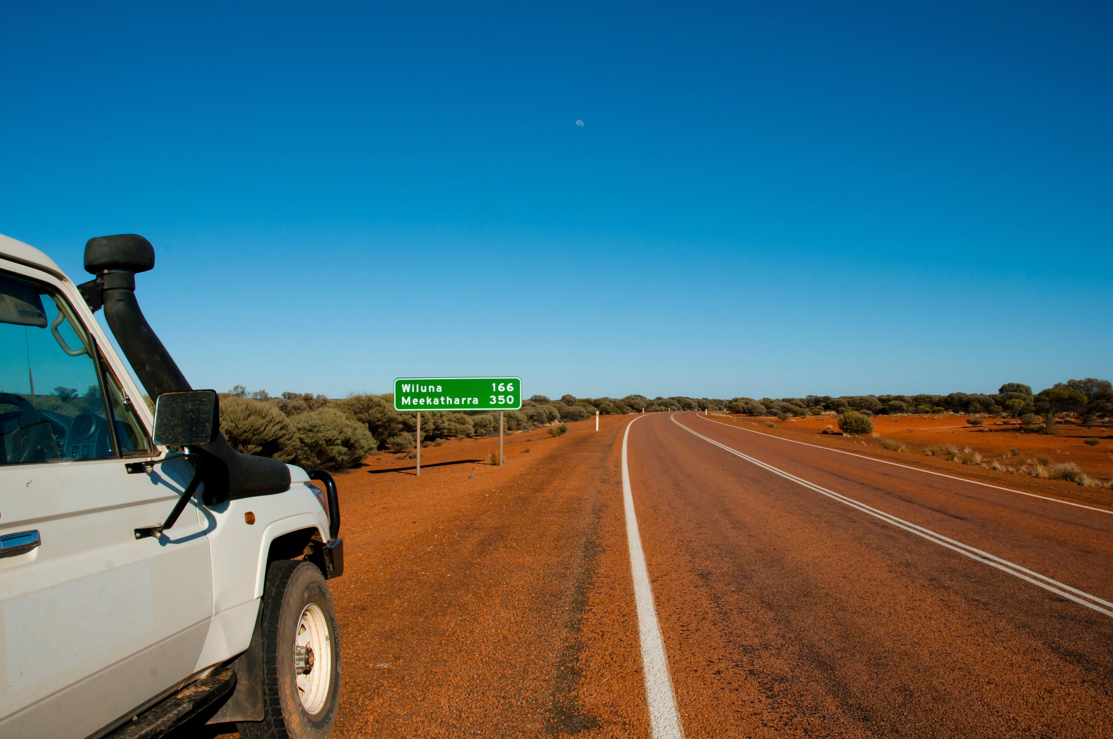 A white vehicle on a red dirt road with a clear blue sky. A green road sign indicates distances to Wiluna and Meekatharra.