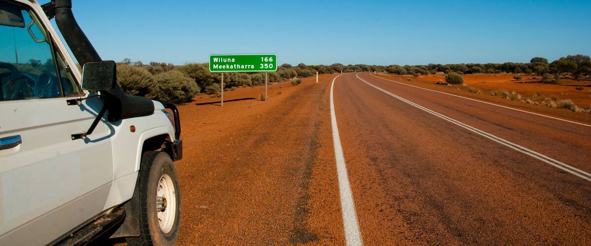 A white vehicle on a red dirt road with a clear blue sky. A green road sign indicates distances to Wiluna and Meekatharra.