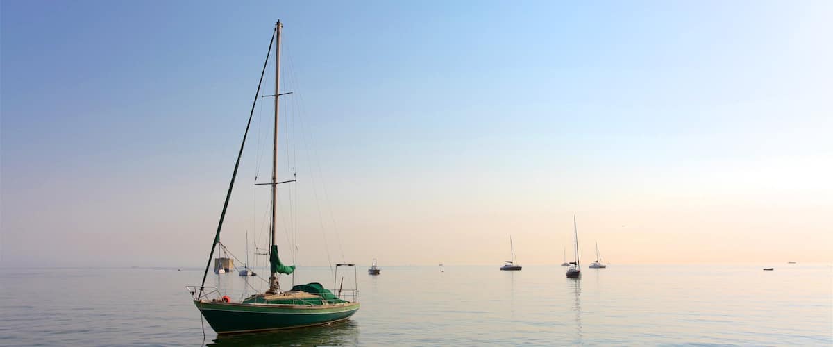 A calm sea with a sailboat anchored in the foreground and several other boats in the distance under a clear sky.