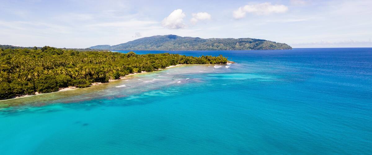 A tropical beach with clear turquoise waters, a sandy shore, and a forested island in the distance.