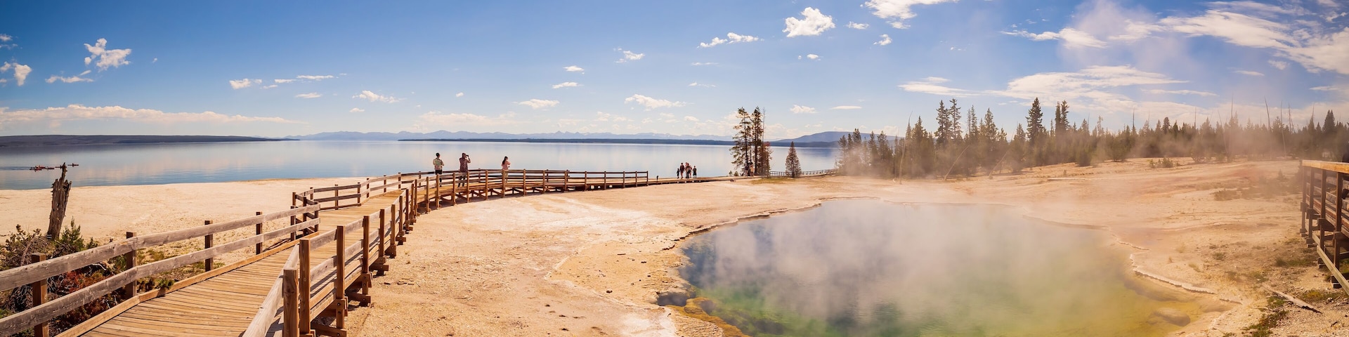Pasarela de madera que conduce a una fuente termal geotérmica, rodeada de árboles y montañas al fondo.