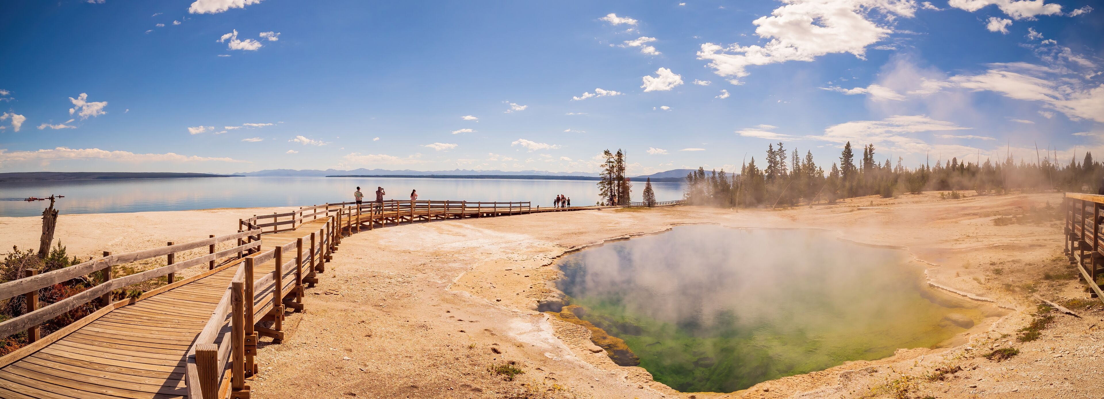 A wooden boardwalk leading to a geothermal hot spring surrounded by trees and mountains in the background.