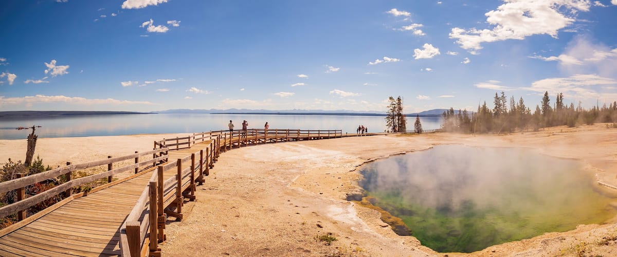 A wooden boardwalk leading to a geothermal hot spring surrounded by trees and mountains in the background.