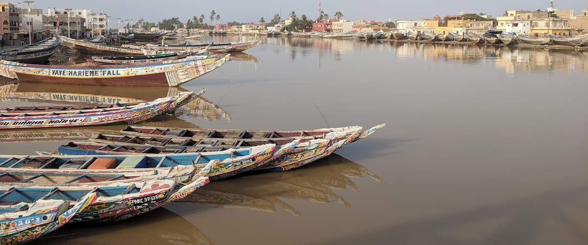 A calm river with several colorful boats docked along the shore. In the background, there are buildings and a mosque.