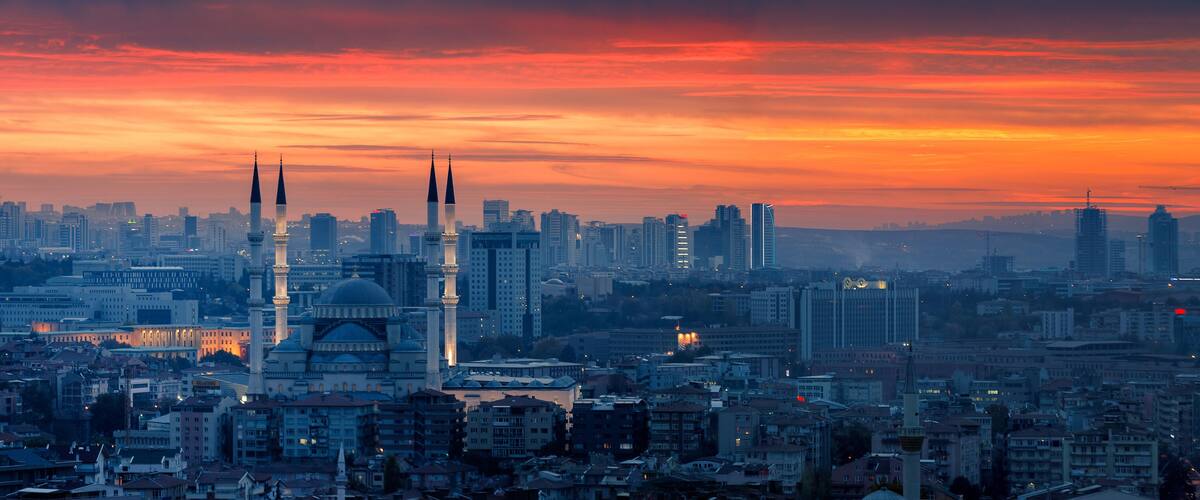 A cityscape at sunset with prominent mosque domes and minarets, surrounded by modern high-rise buildings.