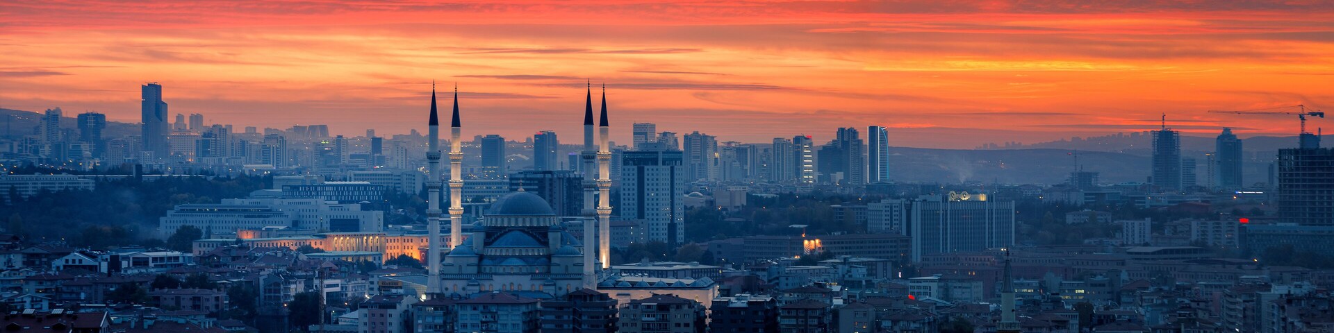 A cityscape at sunset with prominent mosque domes and minarets, surrounded by modern high-rise buildings.