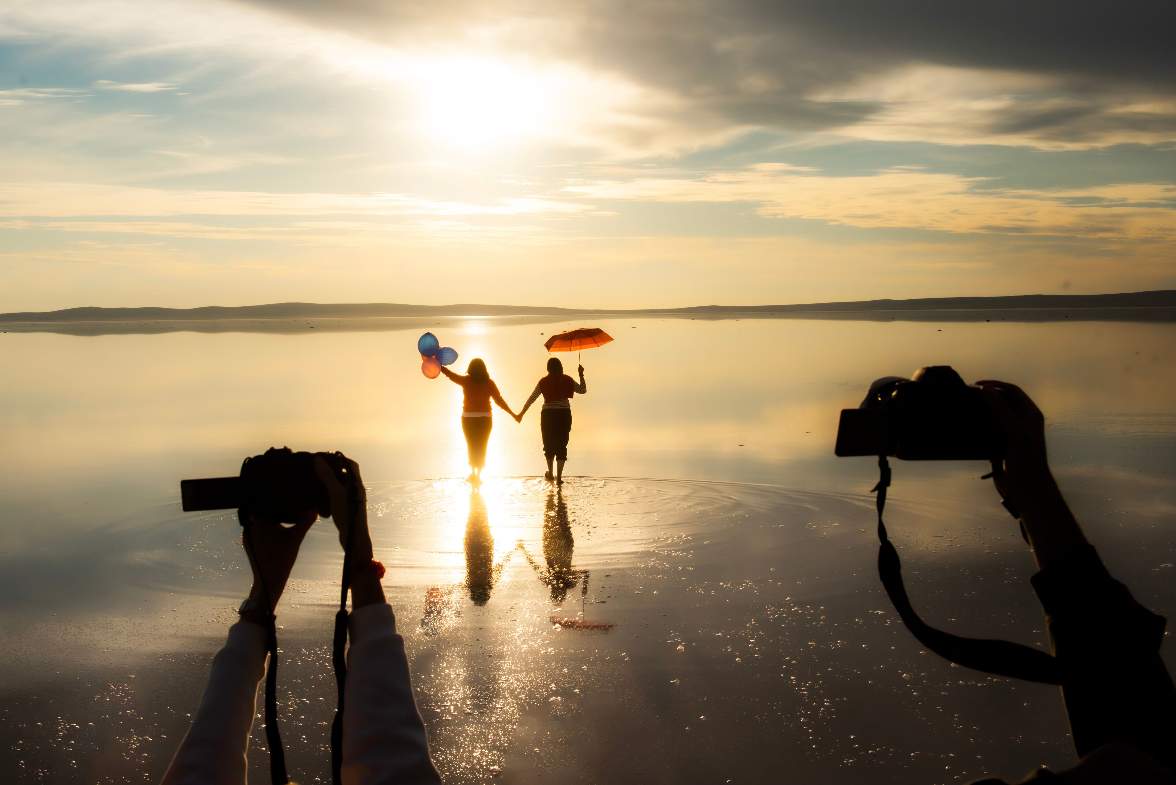 A couple holding hands and balloons on a reflective water surface with a camera capturing the scene.