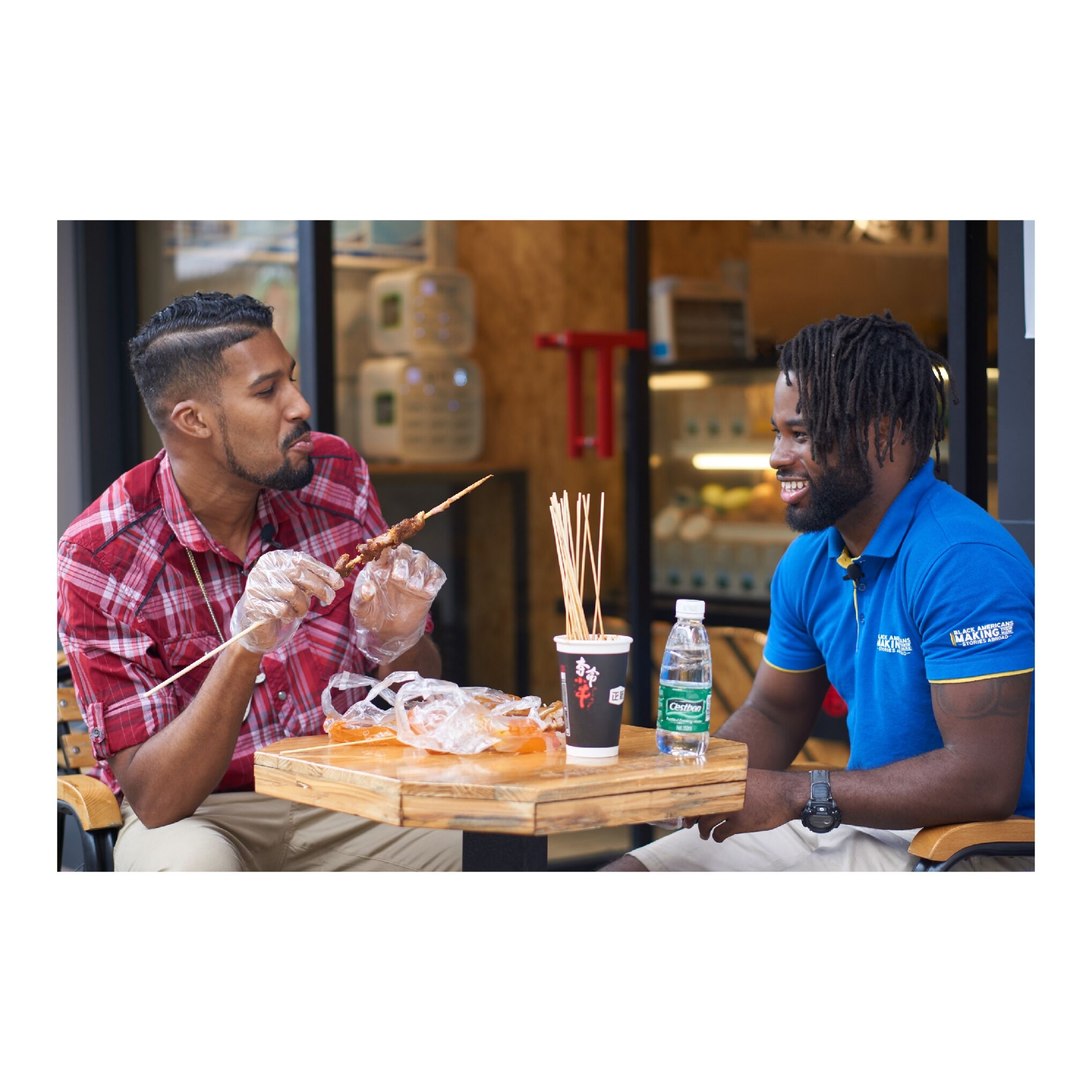 Two people sitting at a table with skewers of food, a drink, and a bottle of water.