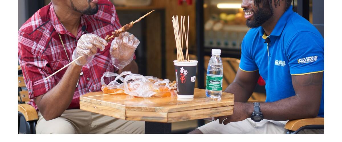 Two people sitting at a table with skewers of food, a drink, and a bottle of water.