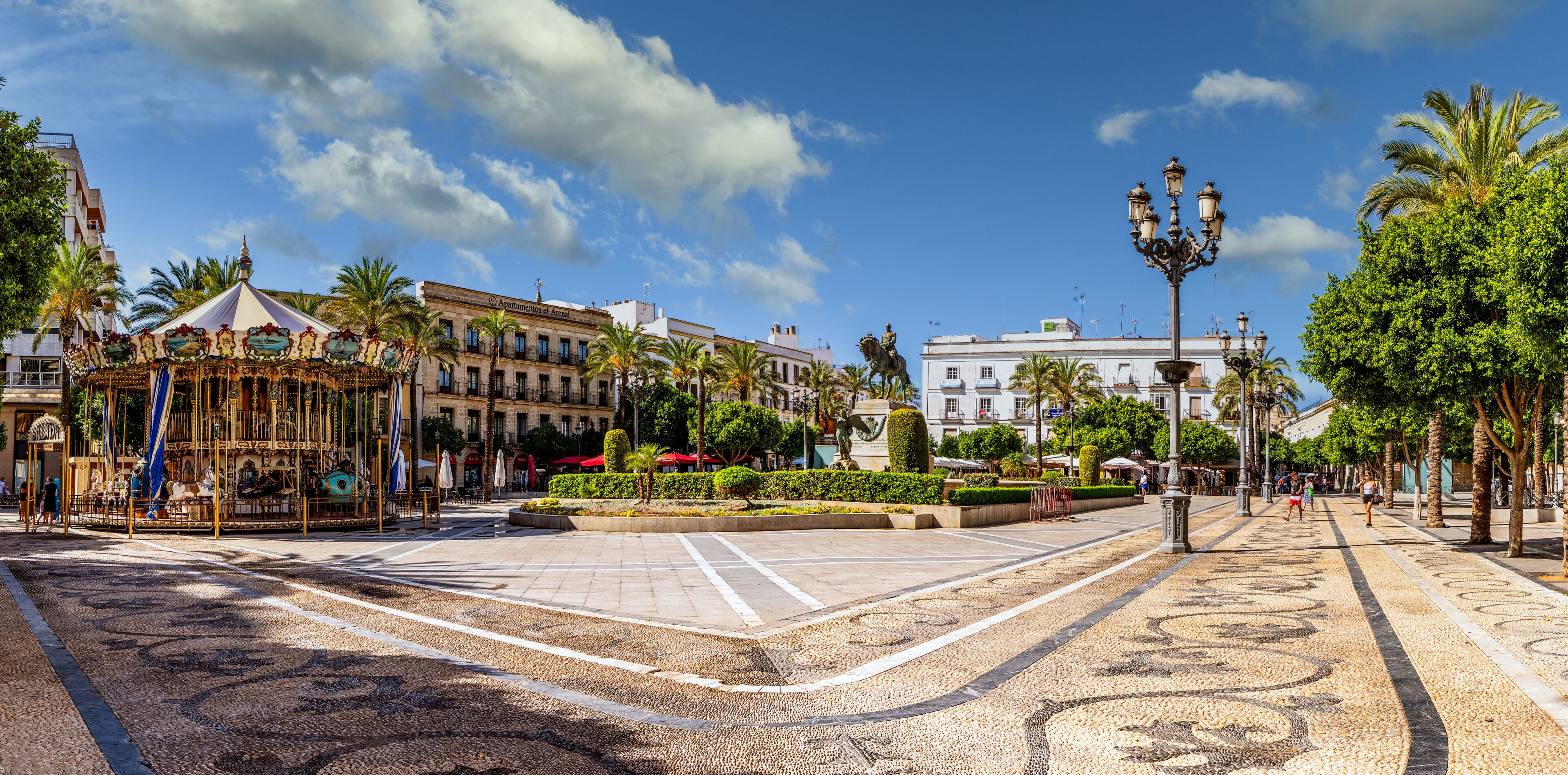 Uma praça com um carrossel, palmeiras e um edifício com a fachada que indica 'Ayuntamiento de Alicante'.