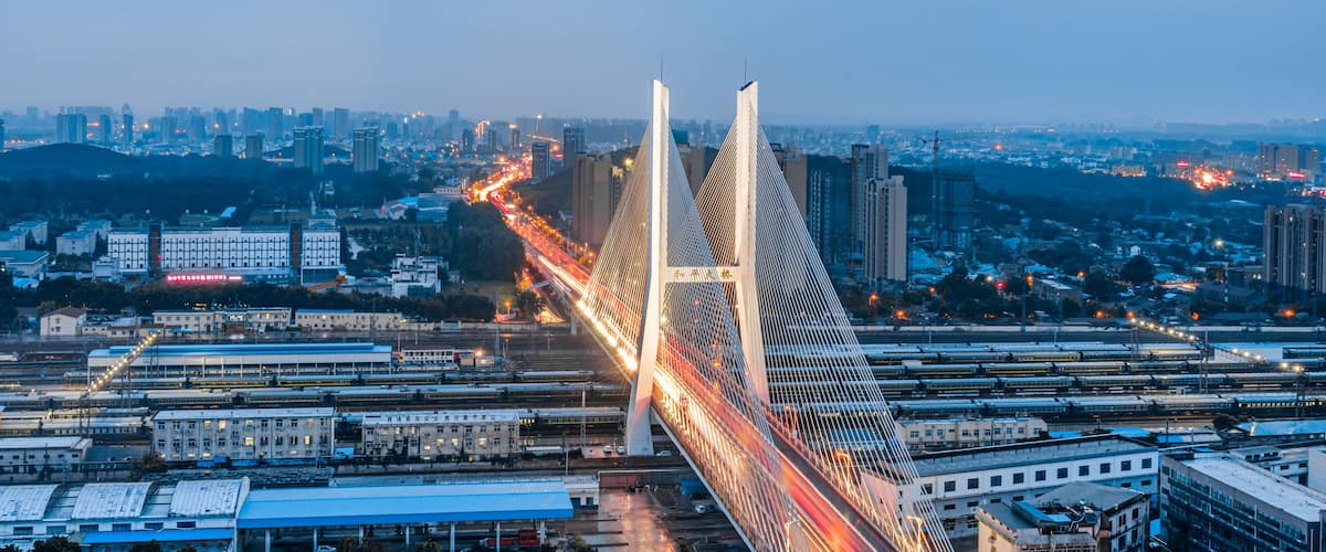 A modern bridge with two tall towers, surrounded by urban buildings and a busy railway track.