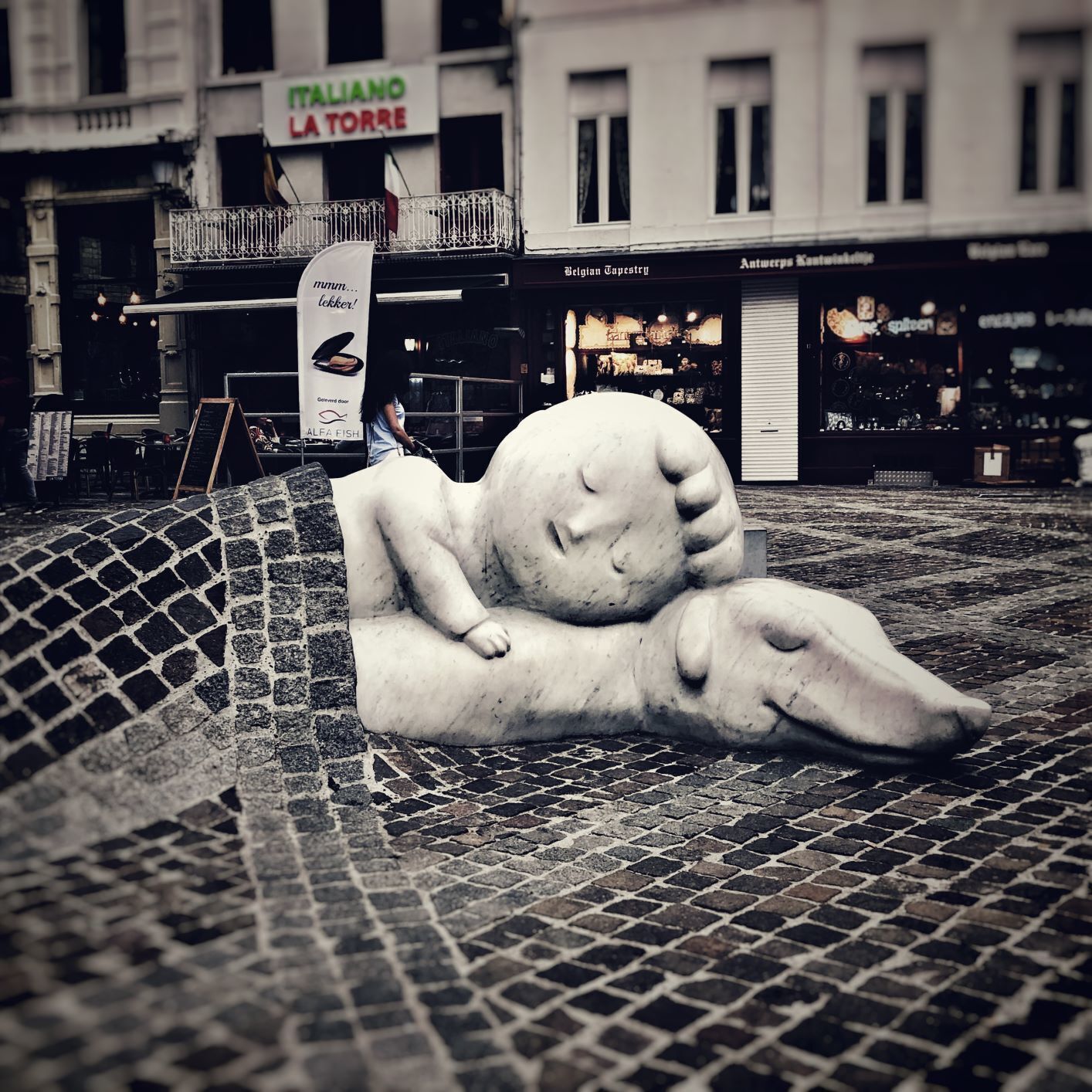 Uma escultura de uma figura reclinada em pavimento de calçada, com uma placa ao fundo indicando 'ITALIANO LA TORRE'.