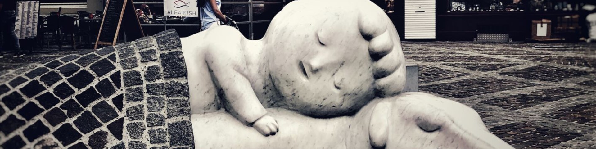 Uma escultura de uma figura reclinada em pavimento de calçada, com uma placa ao fundo indicando 'ITALIANO LA TORRE'.