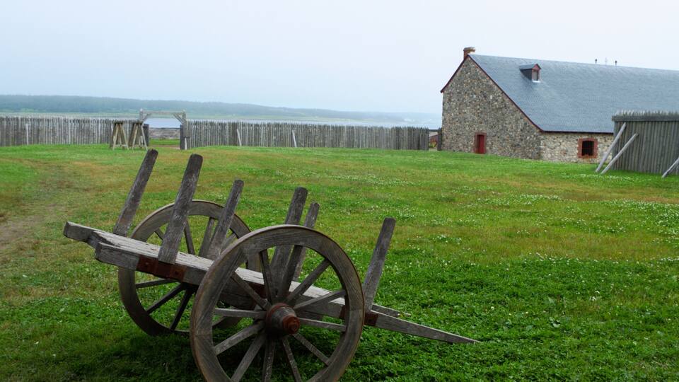 A wooden cart in a grassy field with a stone building and a wooden fence in the background.