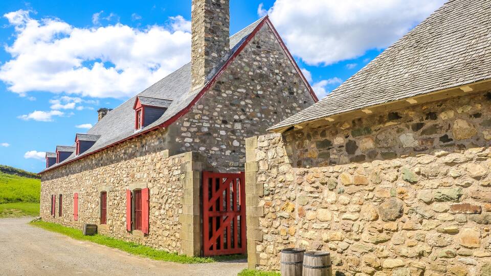 A stone building with a red door and windows, a chimney, and two barrels.