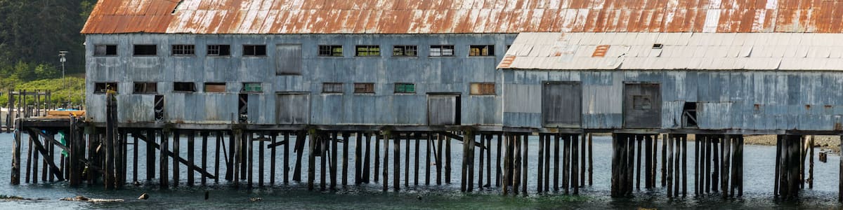 A weathered wooden structure with a rusted roof, situated on stilts over water, surrounded by dense forest.