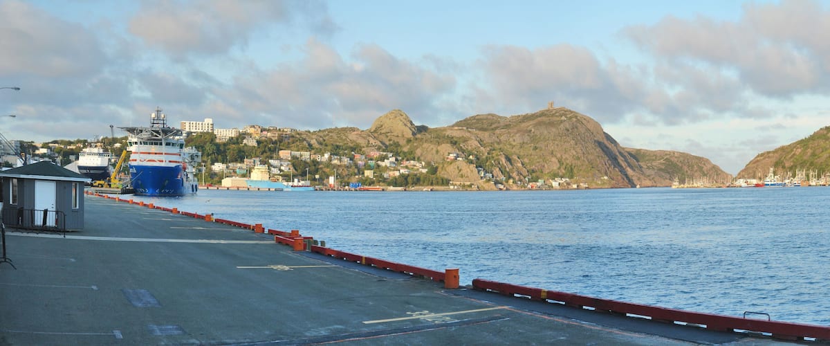 A harbor with a large blue and white ship docked, a red breakwater, and a town with buildings and hills in the background.