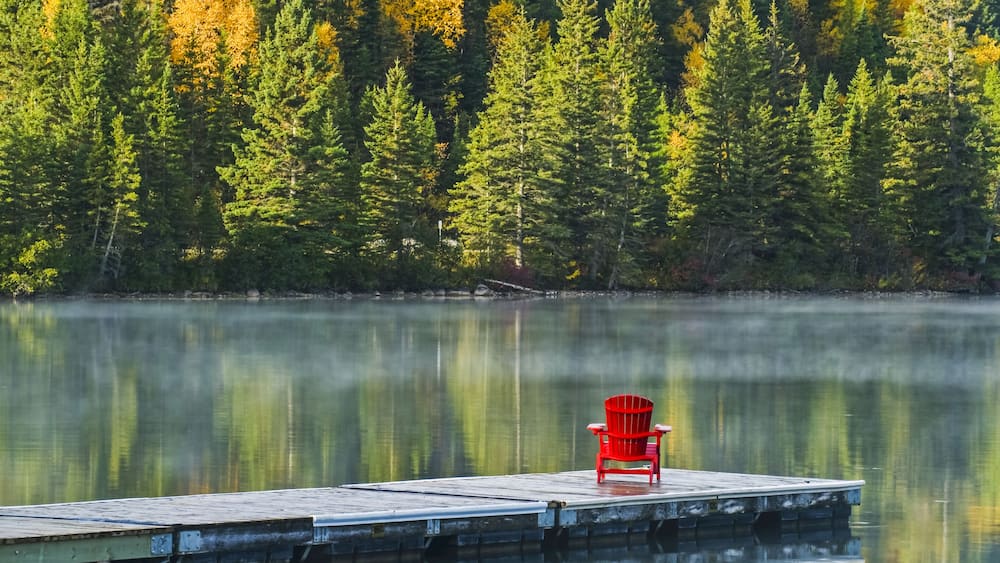 Muskoka chair on dock with autumn coloured foliage reflected in the tranquil lake water of Clear Lake, Riding Mountain National Park; Manitoba, Canada