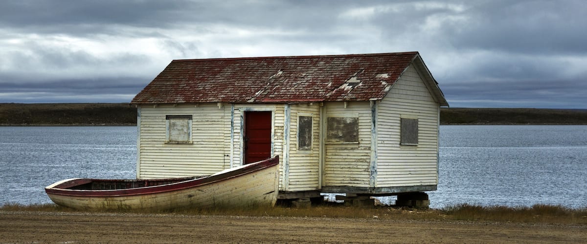 A small, weathered wooden structure with a red door and a boat tied to its side, situated by a body of water under a cloudy sky.