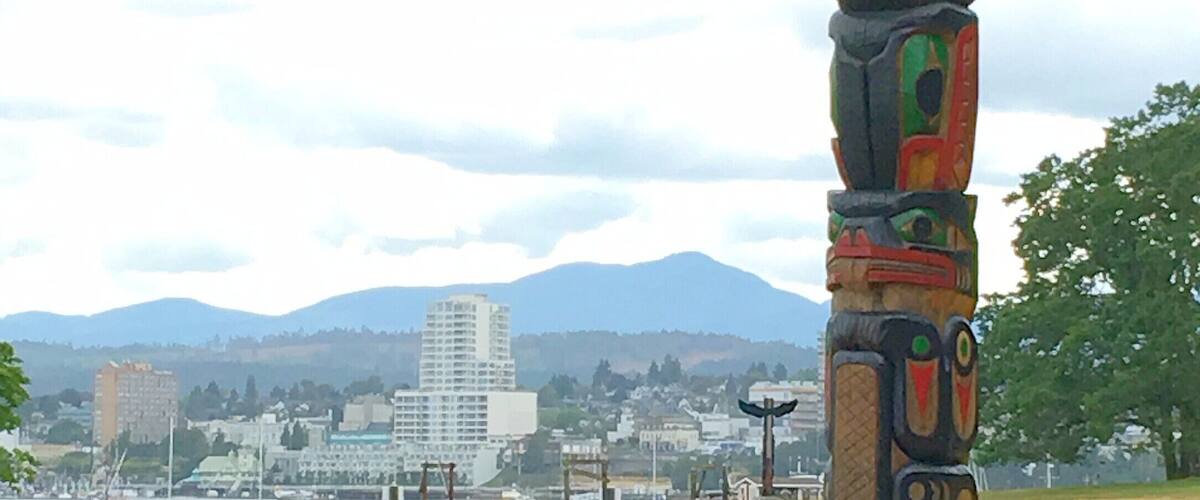 A totem pole in a grassy field with a cityscape and mountains in the background.