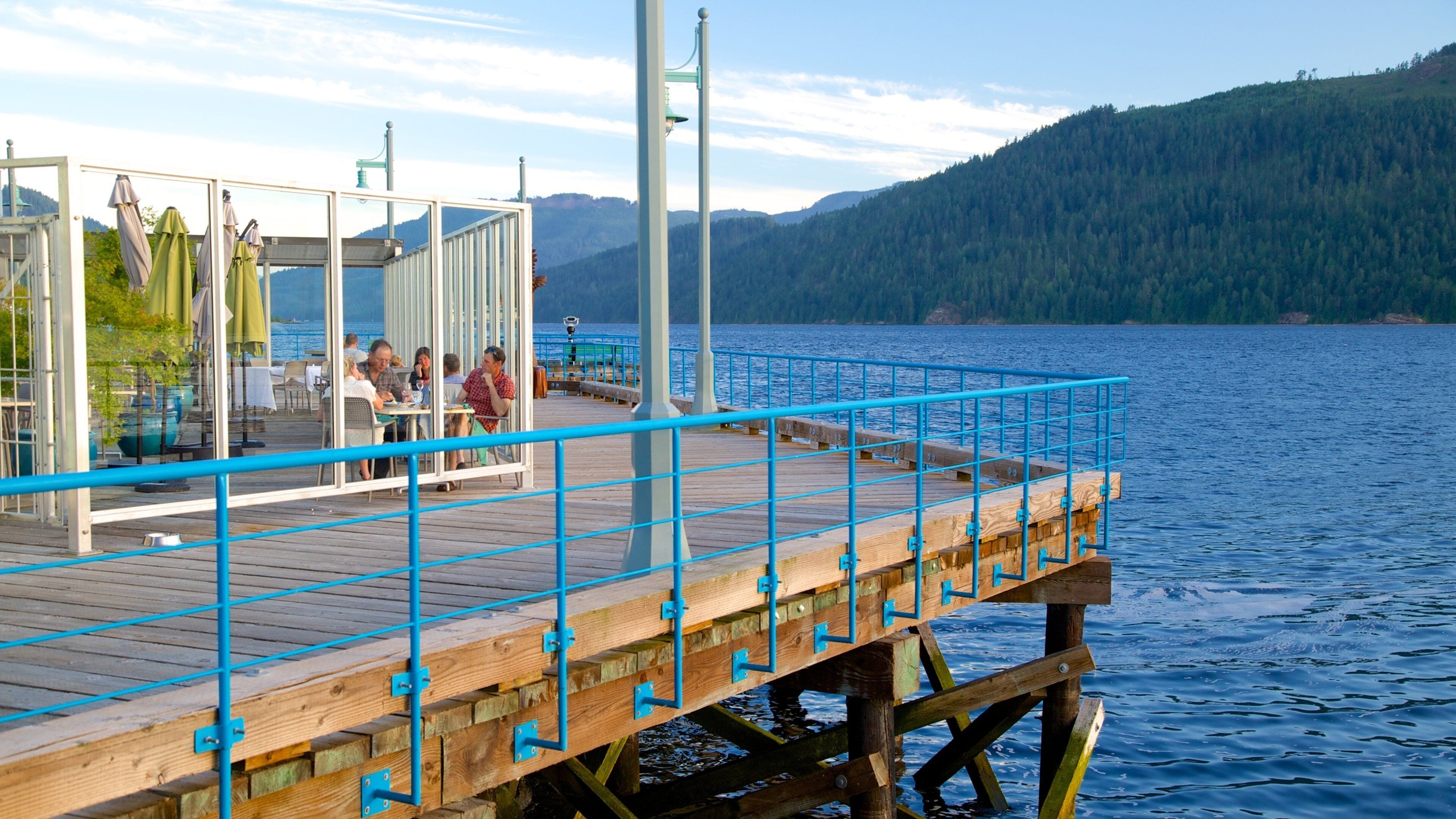 A wooden pier with a blue railing, a covered seating area, and a view of a lake with mountains in the background.
