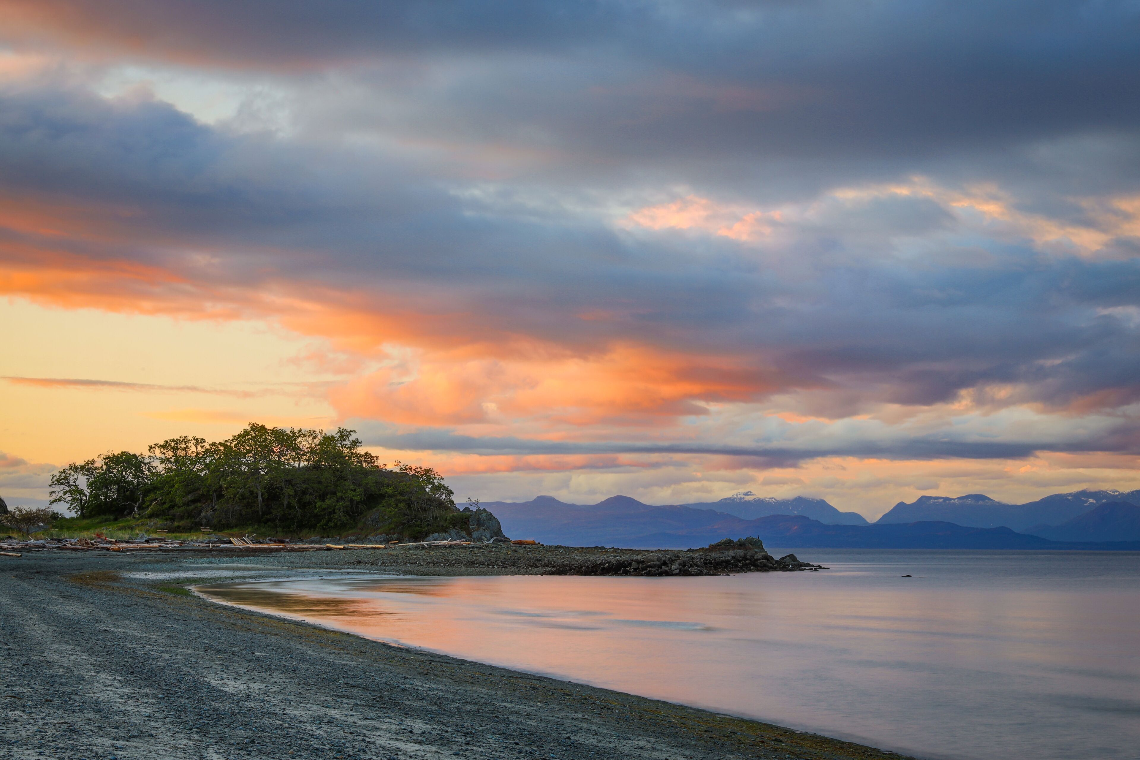 A beach with a calm sea, a rocky shoreline, and a forested island in the distance.