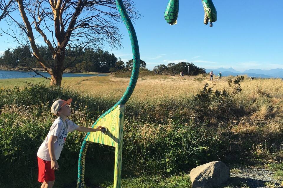 A child interacting with a large, green, snake-shaped sculpture in a park with a lake and mountains in the background.