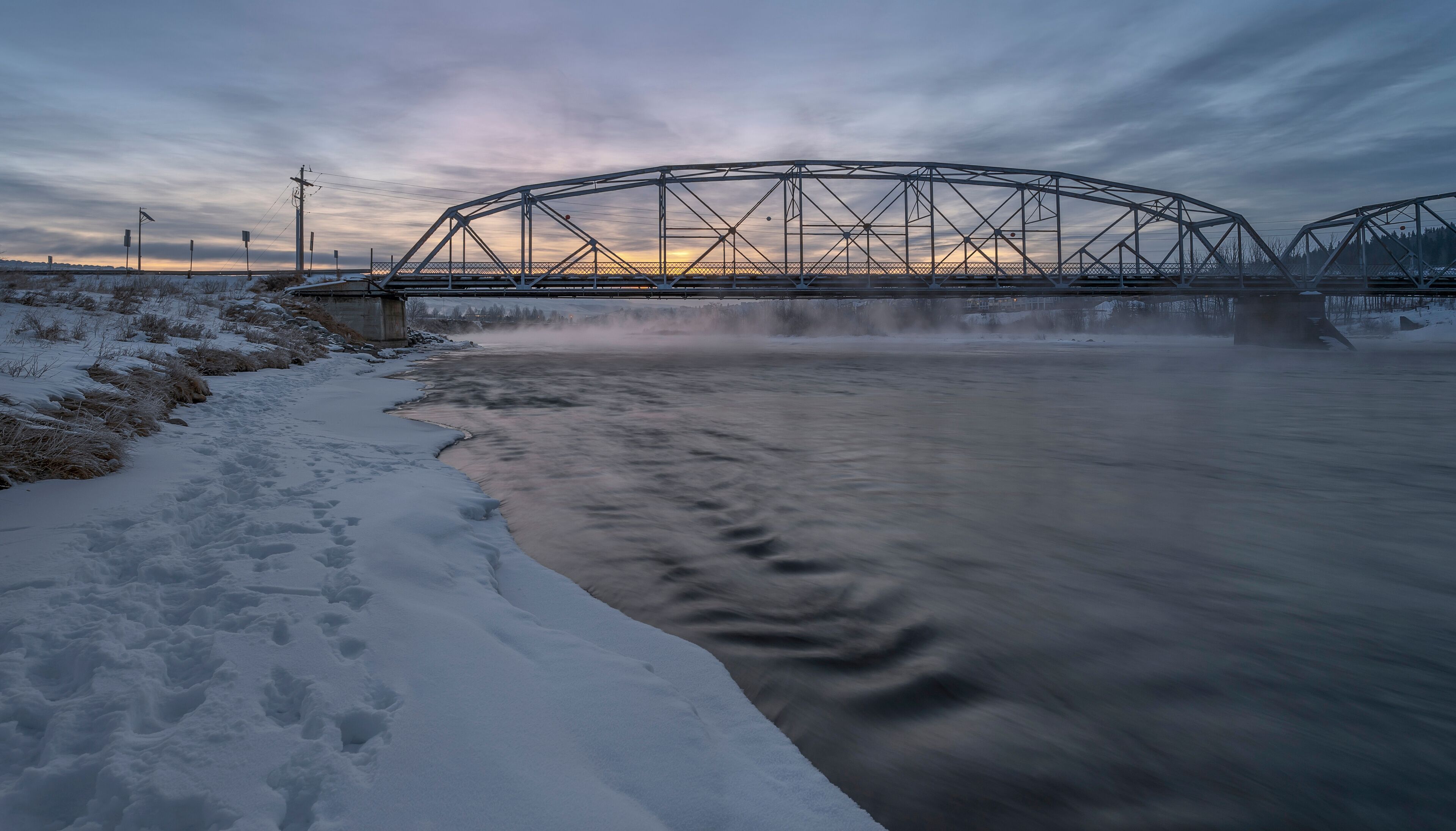 A snow-covered riverbank with a metal bridge in the background.