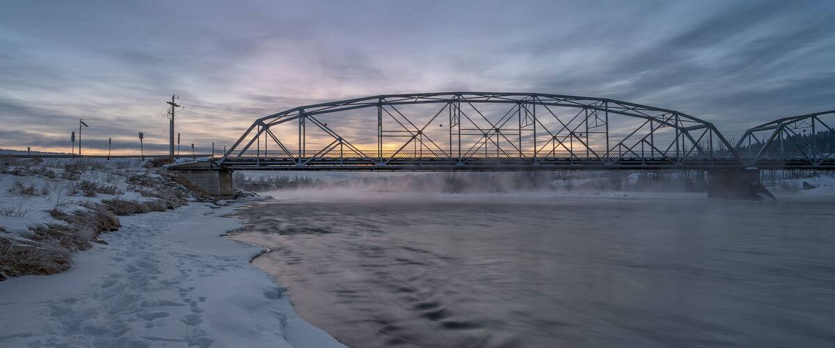 A snow-covered riverbank with a metal bridge in the background.
