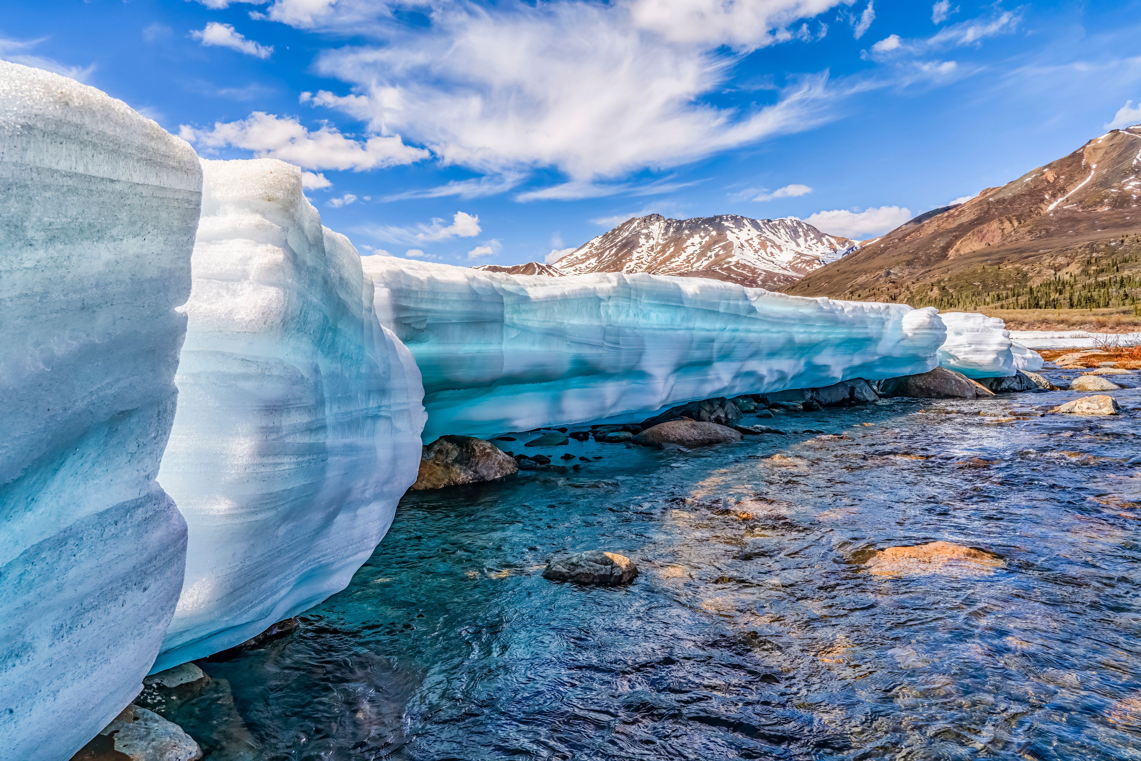 A glacier with a clear blue stream flowing beneath it, surrounded by mountains and a clear sky.