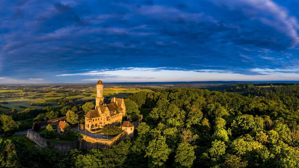 A castle surrounded by dense forest on a hill, with a town in the background and a cloudy sky.
