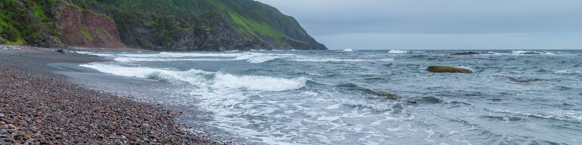 Una playa rocosa con olas rompiendo en la orilla, un acantilado con vegetación verde y un cielo nublado.