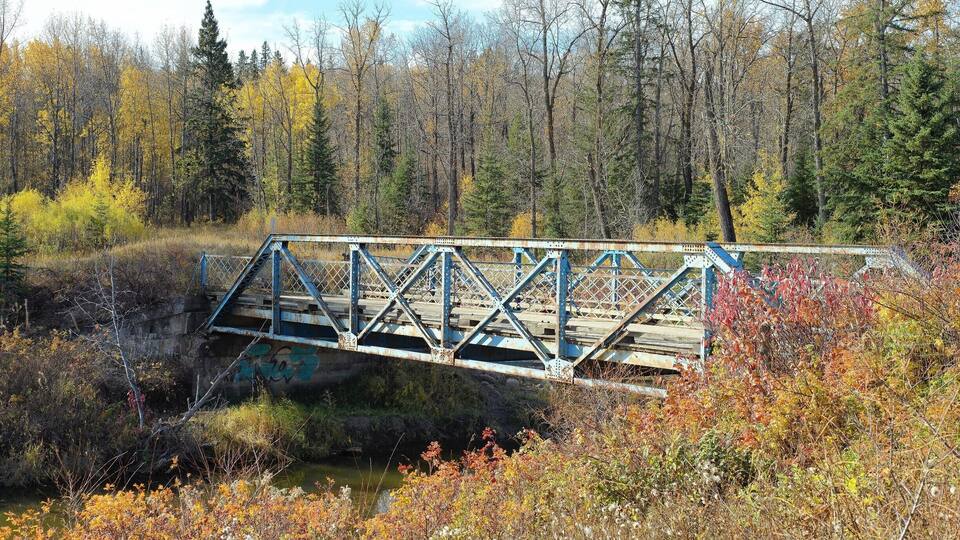 A rusted metal bridge over a river, surrounded by autumn foliage and dense forest.