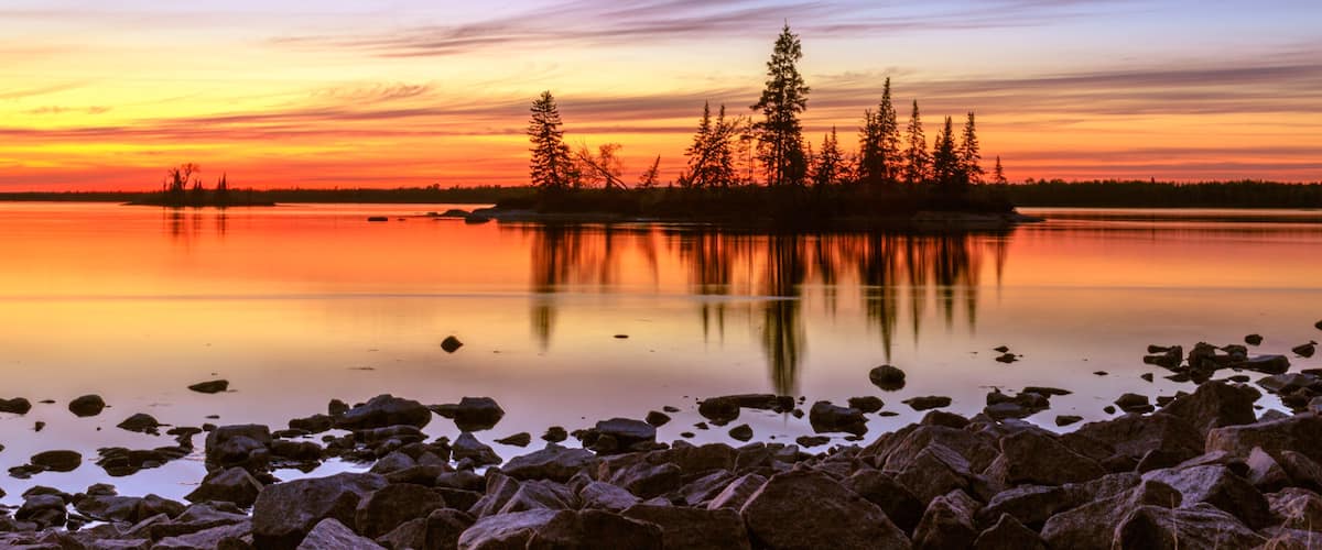 En solnedgång över en stilla sjö med en stenig strand och en grupp träd som avtecknas som silhuetter mot himlen.