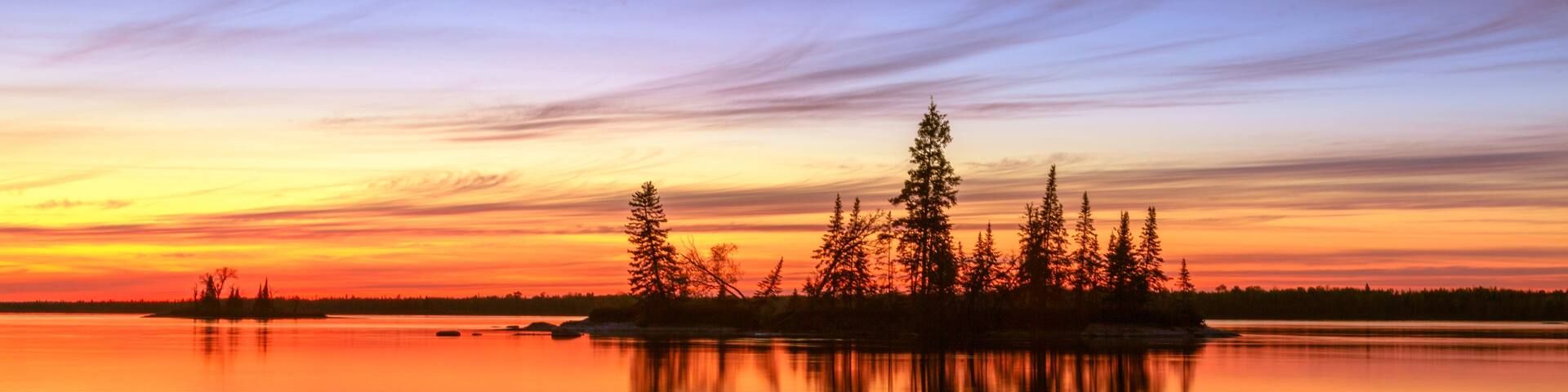 A sunset over a calm lake with a rocky shoreline and a cluster of trees silhouetted against the sky.