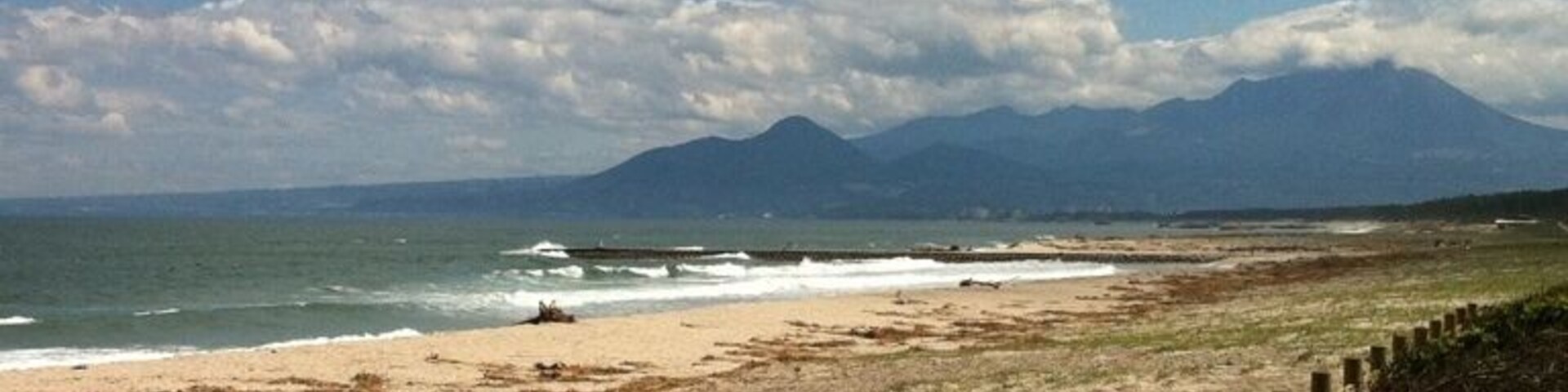 Una playa con una valla de madera y un cartel, olas del océano y montañas a lo lejos.