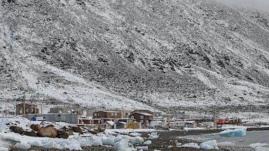 Un paisaje nevado con costa rocosa, edificios y una montaña al fondo.