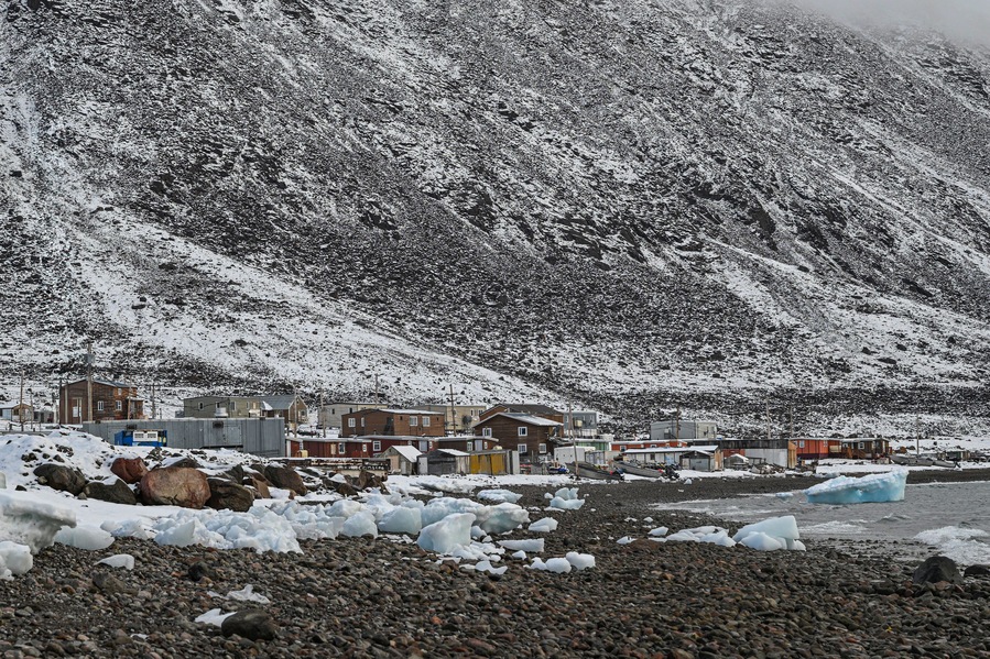 A snowy landscape with a rocky shore, buildings, and a mountain in the background.