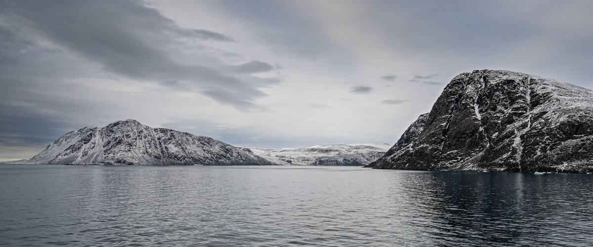 A calm fjord with snow-covered mountains and a cloudy sky.