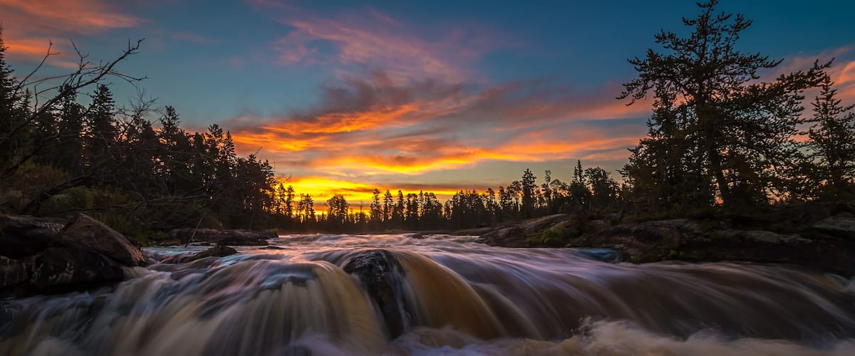 A river flowing through a forest with a sunset in the background.