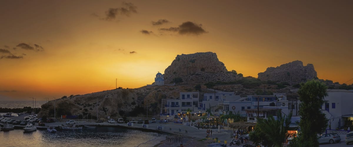 A coastal town at sunset with boats docked, a prominent rock formation, and traditional white buildings.
