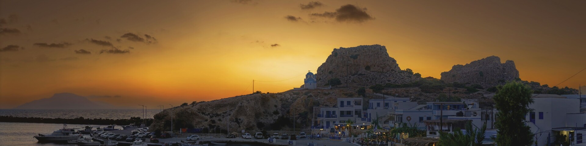 A coastal town at sunset with boats docked, a prominent rock formation, and traditional white buildings.