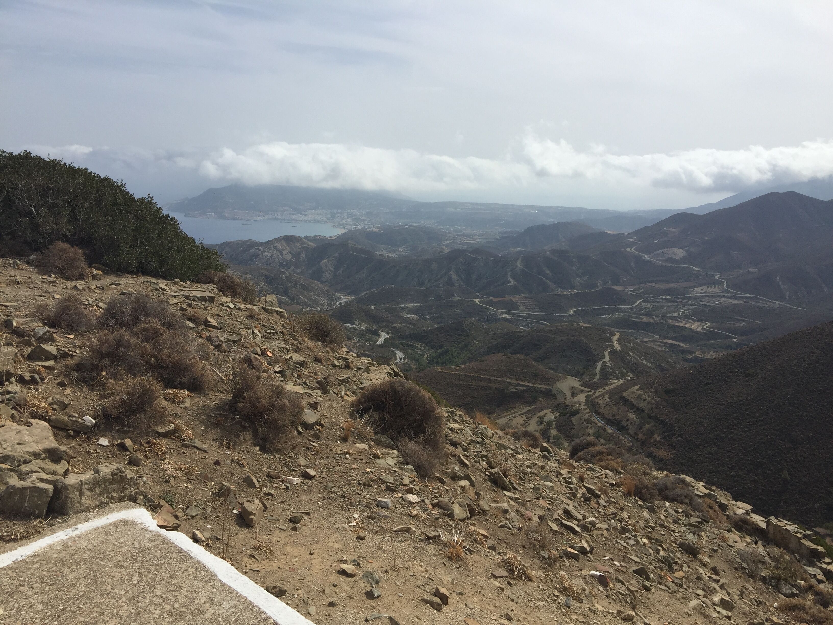 A mountainous landscape with a winding road, rocky terrain, and a distant view of a valley and a body of water.