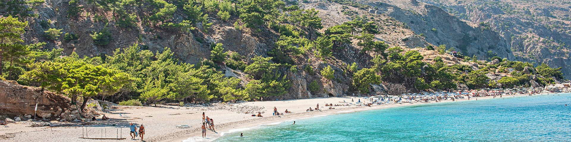 Uma praia turquesa com águas cristalinas, um morro rochoso com vegetação rala e algumas pessoas aproveitando a praia.